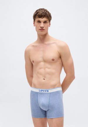 Νέος άνδρας στέκεται γυμνόστηθος φορώντας μπλε μπόξερ της Levi's με λευκή μέση πάνω σε απλό λευκό φόντο.