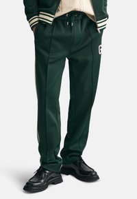 Zielone spodnie dresowe z gładkiego materiału, z elastycznym pasem na sznurek, bocznymi paskami i białym logo po lewej stronie.