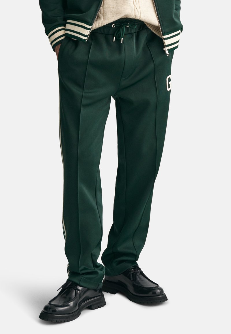 Zielone spodnie dresowe z gładkiego materiału, z elastycznym pasem na sznurek, bocznymi paskami i białym logo po lewej stronie.