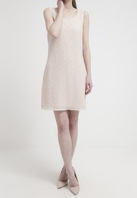 Robe rose courte sans manches avec un motif texturé. Coupe trapèze à longueur genou, associée à des talons de couleur claire. Finition en tissu lisse.