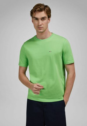 Man die een felgroen T-shirt met korte mouwen draagt, met een klein logo op de borst, gecombineerd met een donkergekleurde broek, staand tegen een grijze achtergrond.