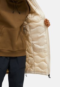 Bruine hoodie met een voorzak, binnen een open beige geïsoleerde jas met een gewatteerde voering en zijritsdetails.