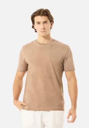 Jeune homme aux cheveux châtain clair portant un t-shirt beige à manches courtes et un pantalon blanc, debout avec une main dans la poche.