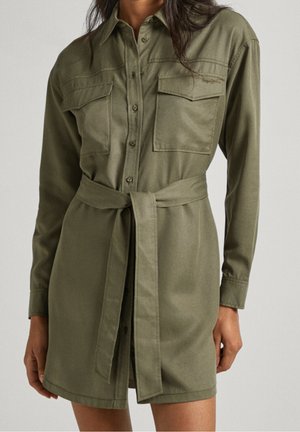 Kobieta ubrana w oliwkowozieloną sukienkę koszulową na guziki z dwoma kieszeniami na piersiach i pasującym paskiem z tego samego materiału związanym w talii.