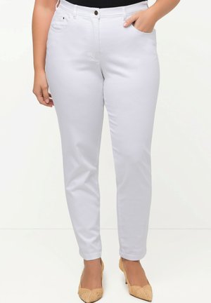Woman wearing fitted white pants with front pockets and beige heeled shoes, standing against a plain light background.