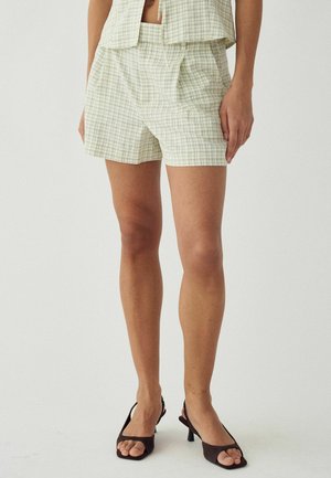 Kobieta ubrana w żółto-zielone szorty w kratę i pasujący bezrękawnik, zestawione z czarnymi sandałami na obcasie z odkrytymi palcami na jednolitym tle.