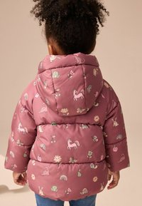 Rosa Puffjacke mit verschiedenen verspielten Mustern, darunter Einhörner und Blumen, mit Kapuze und gestepptem Textil. Blaue Jeans im Denim-Look.