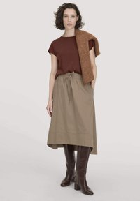 Frau trägt ein braunes kurzärmliges Oberteil, einen beigen Midirock, braune kniehohe Stiefel und einen braunen Strickpullover, der über eine Schulter drapiert ist.