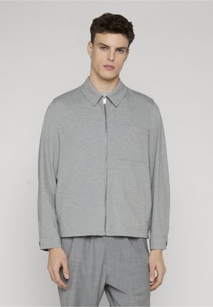 Mandlig model iført en lysegrå lynlåstrøje med brystlomme og matchende grå bukser, stående mod en ensfarvet baggrund.