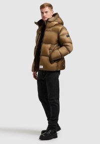 Béžová puffer bunda z nylonu s kapucí, zipovým zapínáním a bočními kapsami. V kombinaci s černými džíny a černými šněrovacími botami.