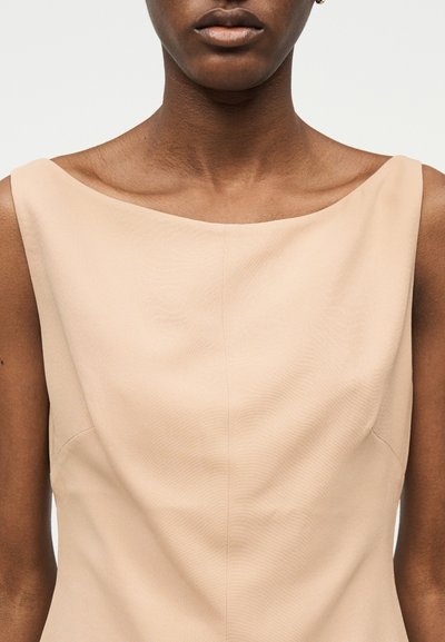 Prikaz izbliza osobe koja nosi bež haljinu bez rukava s vratnim izrezom u obliku broda, na jednobojnoj bijeloj pozadini.