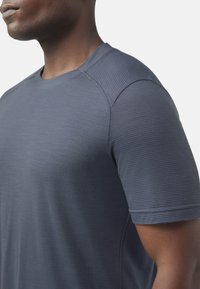 Primo piano di una persona che indossa una maglietta grigia aderente a maniche corte con tessuto testurizzato sulle maniche e sulle spalle, su uno sfondo semplice.