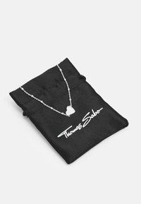 Häpeä sydänkaulakoru ohuella ketjulla, esitelty mustassa nyöripussissa, jossa valkoista "Thomas Sabo" -brändäystä. Pussin pinta on pehmeä.