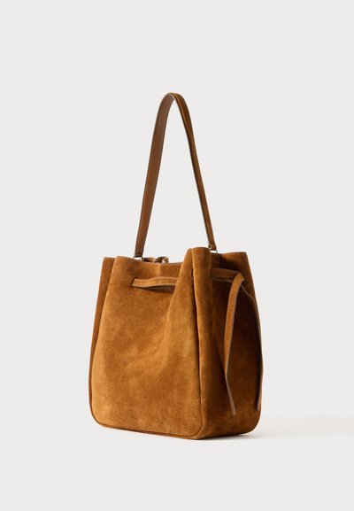Καφέ σουέτ τσάντα tote με δομημένο σχήμα, διαθέτει δύο δερμάτινους ιμάντες και άνοιγμα από πάνω, αναδεικνύοντας απαλή υφή και μινιμαλιστικό σχέδιο.