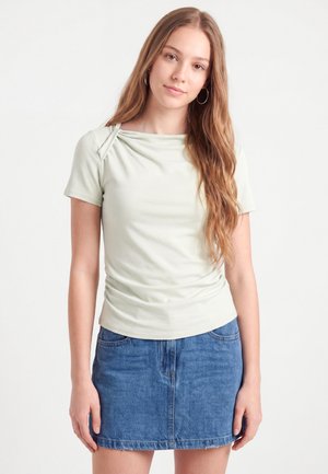 Jeune femme aux longs cheveux châtain clair portant des boucles d'oreilles créoles argentées, un haut à manches courtes vert pâle et une jupe en jean bleue sur fond blanc.