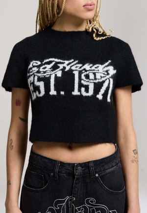 Osoba ubrana w czarny krótki sweter z białym napisem "Ed Hardy Est. 1971" oraz czarne spodnie dżinsowe z wysokim stanem i białym haftowanym wzorem.