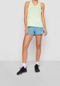 Jasnozielony bezrękawnik, niebieskie szorty, czarno-białe buty sportowe i białe skarpetki z logo, na szarym tle.