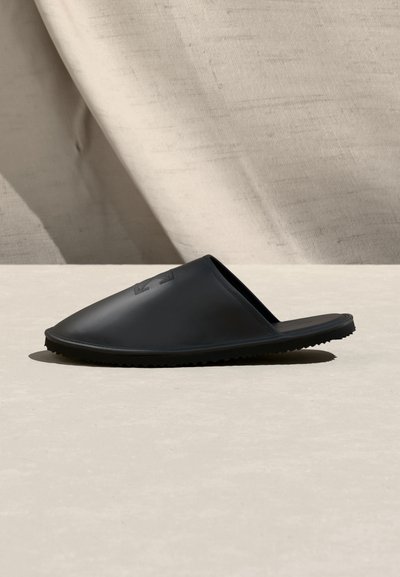 Μαύρο λαστιχένιο σάνδαλο τύπου slide με κλειστό μπροστινό μέρος και υφή στη σόλα, τοποθετημένο σε ουδέτερο μπεζ φόντο.