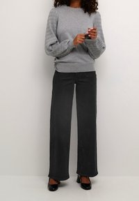 Suéter gris con mangas texturizadas y acanaladas, combinado con unos pantalones vaqueros de pierna ancha negros. Los zapatos de tacón negro con acabado brillante completan el atuendo. Diseño simple y limpio.