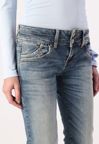 Light blue denim jeans with a slightly faded wash, five pockets, and a double button closure. Features visible stitching and textured fabric.