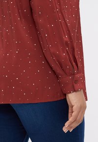 Blouse bordeaux avec des embellissements en sequins argentés, manches longues, poignet à boutons et légèrement froncée aux épaules pour un ajustement décontracté.