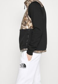 Czarna polarowa kamizelka z brązowym górnym kamuflażem. Posiada zamek błyskawiczny w przedniej kieszeni, wysoki kołnierz i ściągacze przy rękawach. Nosi się ją na białych spodniach.