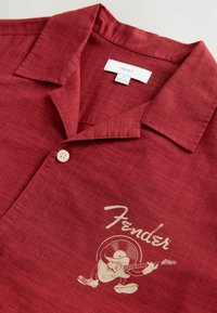 Chemise rouge à boutons avec logo Fender beige et graphique de guitare de dessin animé sur la poitrine gauche, montrant le col et un bouton fermés.