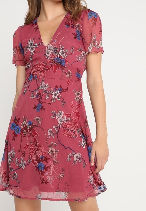Femme portant une robe rose pâle à manches courtes avec un motif floral bleu, blanc et rouge, avec un col en V et une jupe fluide.