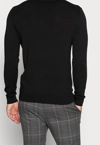 Schwarzer Pullover mit langen Ärmeln, gerippten Bündchen und Saum. Kombiniert mit grauen, karierten Hosen mit braunen Akzenten, die eine figurbetonte Silhouette zeigen.