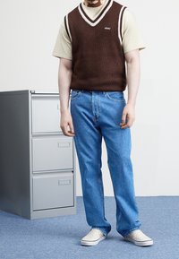 Brown knit vest with white trim over a beige t-shirt, paired with light blue denim jeans and checkerboard slip-on shoes. Gray filing cabinet in background.