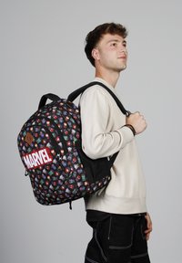 Sac à dos noir avec des motifs colorés de personnages Marvel, grand logo rouge "MARVEL", bretelles rembourrées et poignée supérieure. Tissu léger.