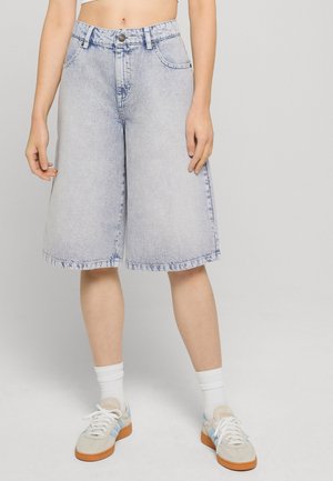 Person iført lyseblå vidde-jeansshorts, hvide sokker og beige sneakers med blå striber, stående mod en ensfarvet baggrund.