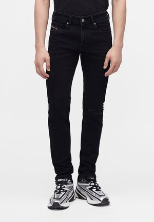 Homme portant un jean slim noir avec une petite déchirure au genou droit et des chaussures de sport argentées et noires, debout les bras détendus.