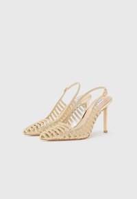 Décolleté slingback in oro con punta affilata, caratterizzate da un design a strisce e una finitura testurizzata. Tacco alto sottile e lati aperti che rivelano la struttura del piede.