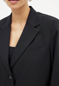 Blazer noir avec un col à revers, fermeture à un bouton et une petite poche poitrine. Fabriqué en tissu texturé avec une coupe ajustée.