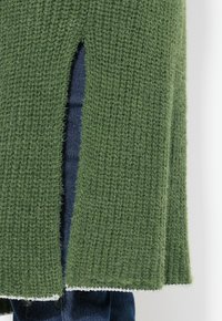 Grön stickad tröja med en texturerad yta, med en sidospalt och vit fåll. Bärs över mörk denim.