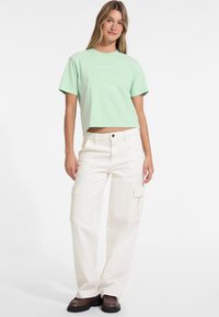 Minttuvihreä lyhythihainen t-paita, jossa on "Guess Jeans" -painatus, yhdistetty valkoisiin cargo-housuihin, joissa on sivutaskut ja rento istuvuus, sekä ruskeisiin kenkiin.