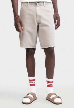 Άνδρας που φοράει μπεζ σορτς μήκους μέχρι το γόνατο, λευκές κάλτσες με κόκκινες ρίγες και γράμματα, και ταμπά σανδάλια με διπλό λουρί, στέκεται σε γκρι φόντο.