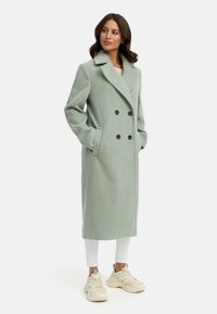 Dubbelzijdige mintgroene wollen jas met grote zwarte knopen, zijzakken, gegoteerde revers en een lange, rechte silhouet.