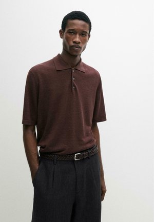 Giovane uomo con indosso una polo marrone a maniche corte, pantaloni grigio scuro e una cintura marrone intrecciata, in piedi contro uno sfondo chiaro uniforme.