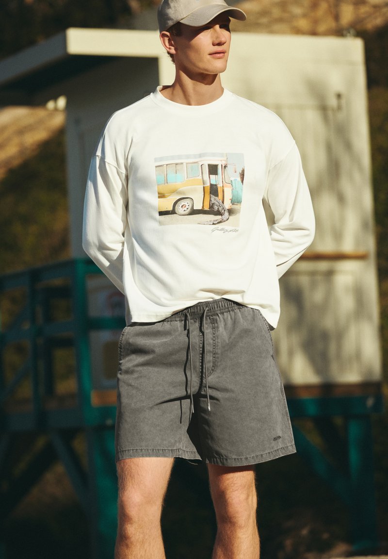 Jeune homme portant une casquette blanche, un sweat-shirt blanc avec un motif de bus et un short gris à cordon, debout à l'extérieur près d'une structure en bois.