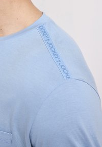 Nærbillede af en lyseblå skjorte skulder med en "Jockey" mærket stribe langs sømmet nær halsudskæringen.