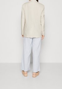 Béžový lněný blazer s přiléhavým střihem, kombinovaný se světlomodrými širokými kalhotami. Oblečení se vyznačuje jednoduchým designem a neutrální barevnou paletou.