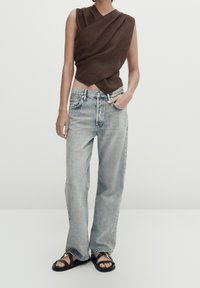 Haut marron sans manches avec un design enveloppé ; jeans bleu clair à coupe ample ; sandales noires ; décor minimaliste avec fond uni.