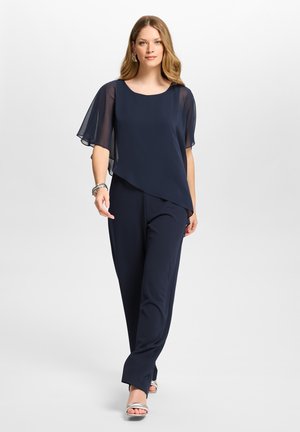 Femme portant un chemisier en chiffon bleu marine à manches courtes avec un ourlet asymétrique et un pantalon large assorti, marchant sur un fond blanc.