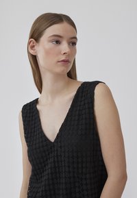 Top noir sans manches avec un décolleté en V. Présente un motif texturé avec de petites découpes circulaires. Tissu lisse avec une coupe ample.