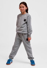 Szara bluza i dopasowane spodnie dresowe, oba z czarnymi łatami z tkaniny. Model nosi niebieskie trampki z białymi akcentami. Gładka faktura.