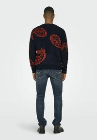 Marineblauwe trui met rood paisleypatroon, ronde halslijn, geribbelde manchetten. Gecombineerd met strakke donkere jeans en zwarte schoenen.