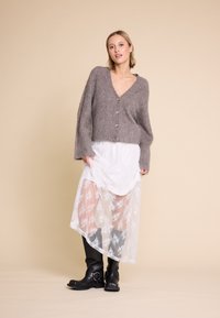 Grå, fuzzy cardigan med V-hals og knapper, lagt over en hvit blomstret lace-kjole, kombinert med knelange svarte støvler.