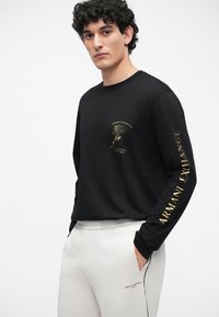 Čierne tričko s dlhými rukávmi a zlatou grafikou "Armani Exchange" na hrudi a rukáve; spárované s svetlosivými nohavicami so značkovými detailmi.